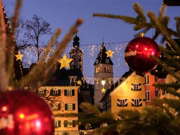 <p>Eine festliche Szene mit Weihnachtsbaumkugeln im Vordergrund und beleuchteten Sternen im Hintergrund. Im Hintergrund ist ein Kirchturm sichtbar, umgeben von dekorierten Häusern.</p>