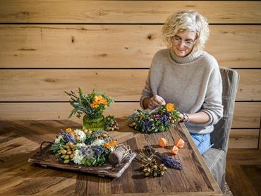 Eine Frau sitzt an einem Holztisch und gestaltet florale Arrangements. Neben ihr befinden sich verschiedene Blumen und Dekorationen.