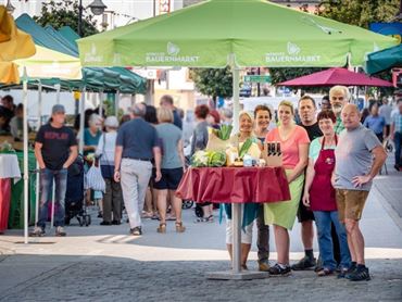 Ein lebhafter Wochenmarkt mit verschiedenen Ständen und vielen Besuchern. Eine Gruppe von Verkäufern lächelt hinter einem roten Tisch mit frischen Produkten.