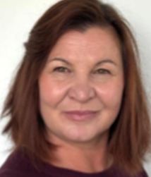 Eine Frau mit dunklem, lockigem Haar lächelt freundlich. Sie trägt eine weiße Bluse und hat ihre Arme verschränkt.