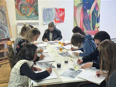 Eine Gruppe von Künstlern sitzt an einem Tisch und zeichnet. Im Hintergrund sind bunte Gemälde zu sehen.