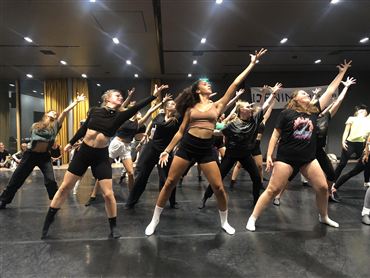 Eine Gruppe von Tänzern übt synchron in einem hellen Raum. Sie tragen Dancewear und zeigen dynamische Bewegungen.