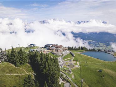 Eine schöne Berglandschaft mit Nebel und einem klaren See. Im Hintergrund sind ein Restaurant und Wanderwege zu sehen.