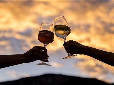 Zwei Gläser werden zum Anstoßen erhoben, silhouettiert vor einem farbenfrohen Sonnenuntergang. Die Szene vermittelt eine festliche und gesellige Stimmung.