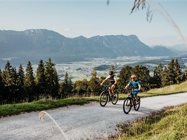 Zwei Radfahrer fahren auf einem Weg in den Bergen. Im Hintergrund sind Täler und Berge zu sehen.
