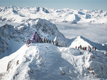Eine beeindruckende Berglandschaft mit schneebedeckten Gipfeln und klarer Sicht. Viele Menschen stehen an einem Gipfelmarker und genießen die Aussicht.