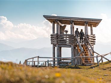 Ein Aussichtspunkt aus Holz mit einer Treppe in den Bergen. Im Hintergrund sind sanfte Hügel und ein blauer Himmel zu sehen.