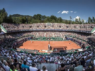Ein Tennisplatz mit Sandbelag und Spielern im Wettkampf. Die Zuschauer sitzen dicht gedrängt in den Tribünen und genießen das Spiel.