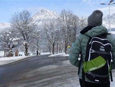 Eine Person steht an einer schneebedeckten Straße und schaut auf die Berge. Umgeben von verschneiten Bäumen, ist die Szenerie ruhig und winterlich.