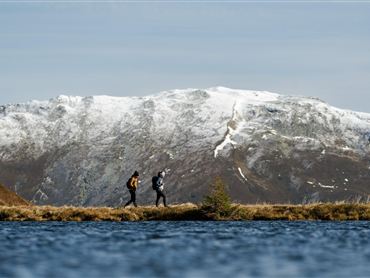 Zwei Wanderer am Ufer eines Sees vor schneebedeckten Bergen. Der Himmel ist klar und die Landschaft ist malerisch.