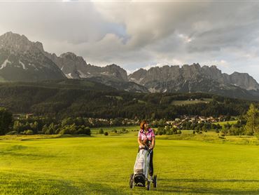Eine Frau mit einem Golfwagen steht auf einem grünen Golfplatz. Im Hintergrund sind beeindruckende Berge und ein bewölkter Himmel zu sehen.