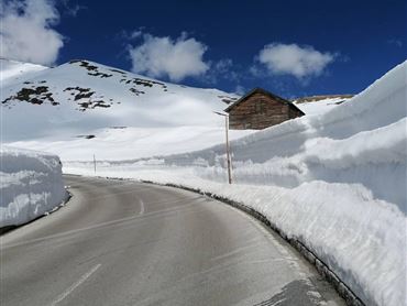 Eine schneebedeckte Straße umgeben von hohen Schneewänden. Im Hintergrund steht ein kleines Holzhaus unter einem blauen Himmel mit einigen Wolken.