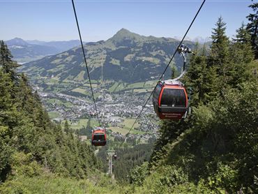 Eine Seilbahn fährt durch eine grüne Landschaft mit Bergen im Hintergrund. Im Tal ist eine Stadt sichtbar.