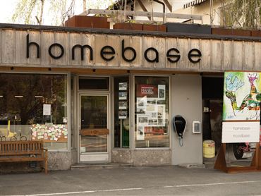 Ein Geschäft mit der Aufschrift "homebase" an der Fassade. Vor dem Laden steht eine Bank, und die Fenster zeigen bescheidene Dekorationen.