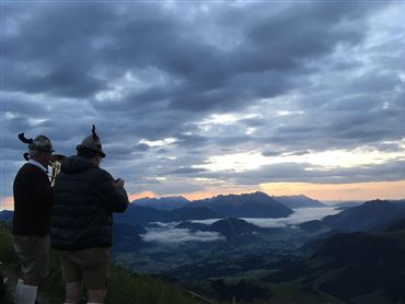 Zwei Männer in traditioneller Kleidung stehen auf einem Berg und betrachten die Landschaft. Der Himmel ist bewölkt und das Licht der Dämmerung strahlt über die Berge.