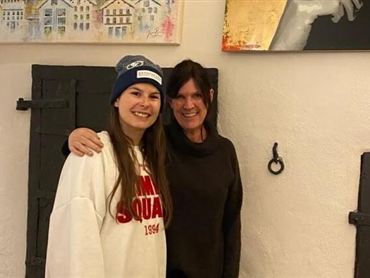 Eine Künstlerin steht vor bunten Gemälden in ihrem Atelier. Im Hintergrund sind kreative Arbeiten und Farben zu sehen.