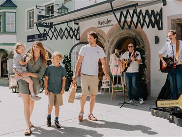Eine Familie genießt einen sonnigen Tag in der Stadt. Im Hintergrund spielen Musiker live Musik.