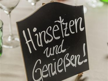 Ein schwarzes Schild mit der Aufschrift "Hinsetzen und Genießen!" steht auf einem Tisch. Um das Schild sind Gläser in verschiedenen Farben angeordnet.