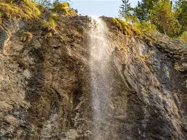 Ein Wasserfall fließt über eine felsige Klippe. Im Hintergrund sind grüne Bäume zu sehen.