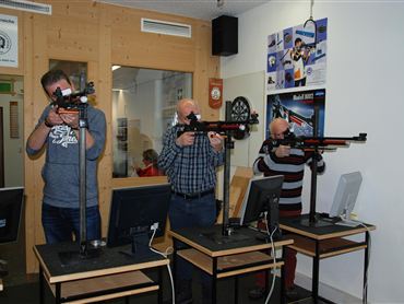 Drei Männer stehen an Schießständen mit Gewehren. Sie konzentrieren sich auf das Zielen und Schießen, während Computer vor ihnen stehen.