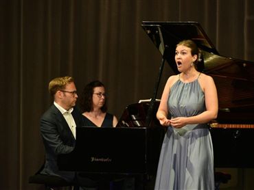 Eine Sängerin trägt ein elegantes Kleid und steht vor einem Klavier. Der Pianist begleitet sie, während eine weitere Person im Hintergrund zuhört.