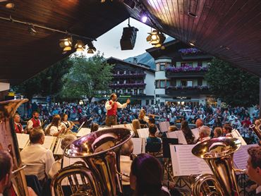 Ein Open-Air-Konzert mit einem Orchester auf einer Bühne. Viele Zuschauer genießen die Aufführung im Freien.