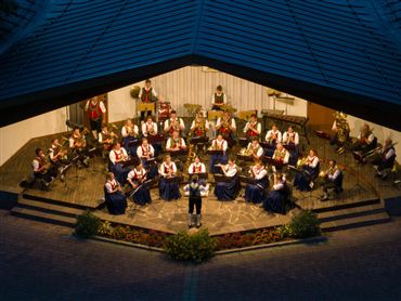 Ein Blasorchester spielt im Freien. Die Musiker tragen traditionelle Trachten und stehen auf einer Bühne.