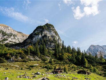 Eine malerische Berglandschaft mit hohen Felsen und saftigem Grün. Im Vordergrund sind einfache Hütten und Nadelbäume zu sehen.