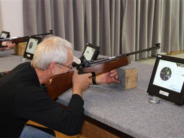 Ein älterer Mann zielt mit einem Luftgewehr auf eine Zielscheibe. Im Hintergrund sind weitere Personen und Schießstand-Ausrüstung zu sehen.