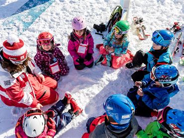 Eine Gruppe von Kindern sitzt im Schnee und hört einem Skilehrer zu. Rundherum sind Skiausrüstungen und verschneite Berge sichtbar.