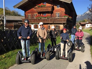 Eine Gruppe von sechs Personen fährt auf Segways in ländlicher Umgebung. Im Hintergrund ist ein traditionelles Holzhaupthaus mit Blumen zu sehen.