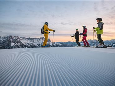 Eine Gruppe von Skifahrern steht auf einer verschneiten Piste mit Bergen im Hintergrund. Der Himmel ist klar und zeigt schöne Farben beim Sonnenuntergang.