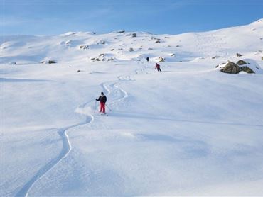 Zwei Skifahrer fahren durch tiefen, frischen Schnee in einer weiten, winterlichen Landschaft. Der Himmel ist klar und blau.