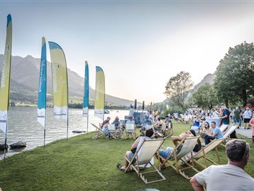 Das Bild zeigt Menschen, die an einem Seeufer in Liegestühlen entspannen und eine Veranstaltung im Freien genießen. Die Atmosphäre wirkt sommerlich und gemütlich, mit Blick auf den See, die Berge und zahlreiche Besucher. 