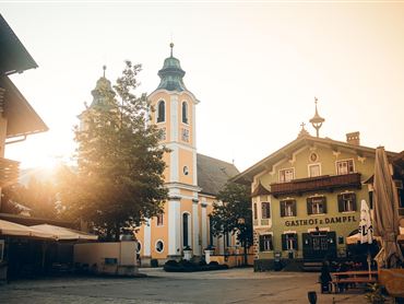 Ein malerischer Dorfplatz mit einer schönen Kirche und einem Gasthof. Die Sonne scheint warm auf die Gebäude, die im traditionellen Stil gehalten sind.