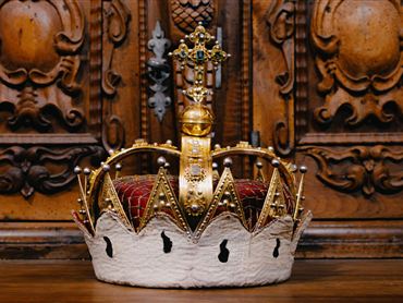 Eine prächtige Krone mit goldenen und roten Elementen steht vor einer kunstvoll geschnitzten Holzfläche. Sie symbolisiert Macht und Königlichkeit.