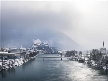 Eine verschneite Stadt am Ufer eines Flusses. Im Hintergrund sind Berge und grauer Himmel zu sehen.