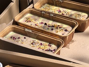 Drei Formen mit Seife, garniert mit getrockneten Blüten, liegen in einem Karton. Die Seife hat eine cremige Textur und eine helle Farbe.