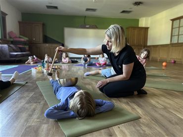 Eine Lehrerin führt eine Entspannungsübung mit Kindern durch. Die Kinder liegen auf Matten, während die Lehrerin eine Schüssel mit einem Löffel bearbeitet.