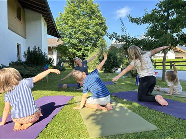 Eine Gruppe von Kindern macht Yoga im Freien auf grünen Matten. Im Hintergrund sind ein Haus und Bäume zu sehen.