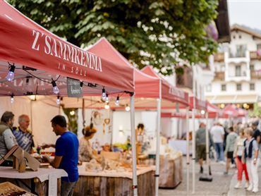 Ein lebendiger Marktplatz bei Dämmerung mit Ständen, die lokale Produkte anbieten. Menschen schlendern durch die Gassen und genießen die Atmosphäre.